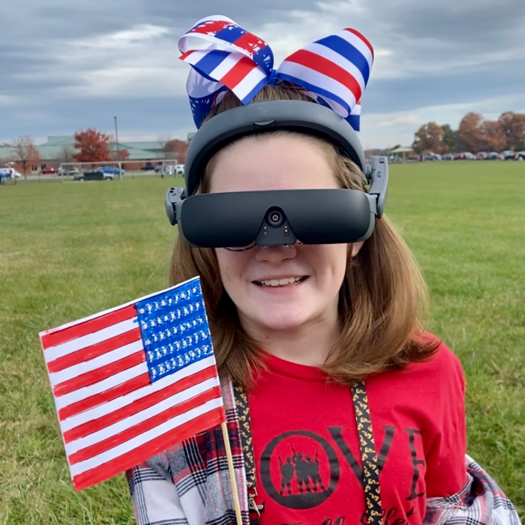 eSight user, 12 year old Rhylee smiles at the camera while wearing the eSight 4 and holding the American flag.